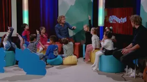 Een foto van een groep kinderen en twee mannen. Ze zitten in een studio. Sommige van de kinderen steken hun vinger op. Een van de mannen wijst naar een kind. Op de achtergrond zie je de tekst "apennoten".
