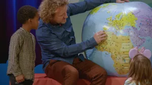 Een foto van een man met een grote opblaasbal in de vorm van de Aarde. Bij hem zitten twee kinderen. De man wijst naar het land Marokko op die wereldbal.