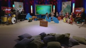 Een foto van een Tv-studio. Rechts zie je een groep jonge kinderen en twee mannen. Een van die mannen speelt op zijn gitaar. Links zie je een band die muziek maakt. Op de voorgrond zie je allemaal kussens. Op de achtergrond staat een groot beeldscherm. 