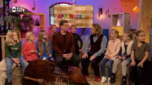 Foto van kinderen en twee mannen die bij elkaar zitten in studioachtige ruimte, op de grond ligt een contrabas.