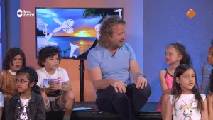 Een foto van presentator Pepijn Gunneweg in een studio. Hij zit tussen kinderen en vertelt hen iets. Op de achtergrond zie je een TV met een tropische foto erop.