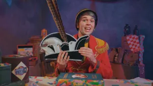 Een foto van een presentator in rood met lichtbruine carnavalskleding en een bruine hoed op achter een tafel. Hij heeft een zwart-witte hoed, een steek genoemd vast. Het is een hoed met een grote veer erop en aan de zijkant vijf zilverkleurige belletjes. 