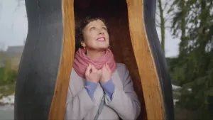 Een foto van een vrouw. Ze staat ergens in. Het lijkt op een holle boom, maar het is een kunstwerk met gladde vormen. Ze houdt haar gebalde handen voor haar borst en kijkt schuin omhoog naar buiten. Op de achtergrond zie je bomen.