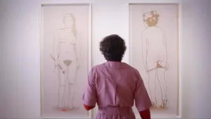 Een foto van een vrouw. Ze heeft een paarse jurk aan en je ziet haar op de rug. Ze kijkt naar twee grote tekeningen van blote vrouwen. Die tekeningen hangen aan een witte muur. 