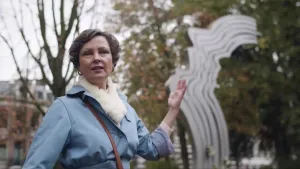 Een foto van presentator Wieteke die buiten staat met een opgestoken hand. Achter haar opgestoken hand is een kunstwerk zichtbaar die lijkt op de zijkant van meerdere gezichten die naar boven kijken.