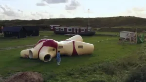 Een foto van presentator Wieteke van ver af. Ze loopt naast een kunstwerk van een vrouwelijk lichaam zonder een hoofd, armen en benen. Het kunstwerk is hol van binnen en er zitten verschillende gaten in waardoor je naar binnen kan kijken.