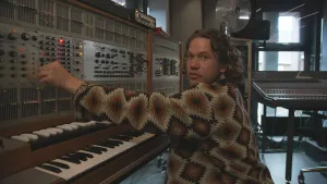 Een foto van Pascal die achter een synthesizer zit, een soort elektronische piano. Daarboven zitten draaiknopjes, schuifjes en lichtjes. Pascal draait aan één van de knopjes. Met de synthesizer kan elektronische muziek met geluidseffecten gemaakt worden.