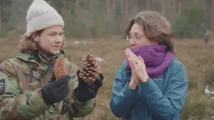 Een foto van een grasvlakte in een bos. Pascal staat met een muts op naast Annemarit van Broekhoven, die een sjaal omheeft. Pascal heeft twee grote dennenappels vast in zijn gehandschoende handen. Annemarit heeft haar handen gevouwen tegen elkaar aan. 