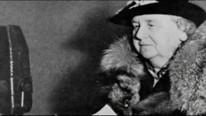 Een foto van koningin Wilhelmina die rechts in beeld is. Links in beeld staat een grote, zwarte microfoon. Wilhelmina heeft een bondkraag om haar nek en een zwarte hoed op. Ze spreekt tijdens de Tweede Wereldoorlog op de Radiozender BBC in Londen.