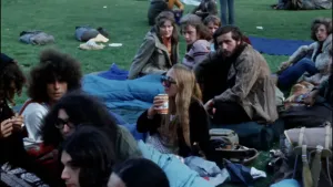 Een foto van mensen op een grasveld in de zomer in het vondelpark in Amsterdam. De mensen zitten op het gras op een blauw kleed. Er zitten ook twee mensen in een lichtblauwe slaapzak. Eén vrouw drinkt uit een blikje. Er liggen ook rugzakken op de grond.