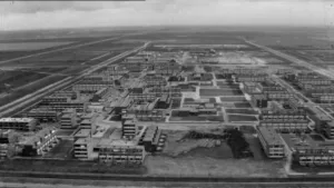 Een zwart-wit foto van de stad Lelystad. De foto is vanuit de lucht genomen. Er zijn op de foto allemaal gebouwen te zien van meer dan één verdieping met straten ertussen. Op de achtergrond ligt er ook nog ongebruikte, rechthoekige stukken natuurgrond. 