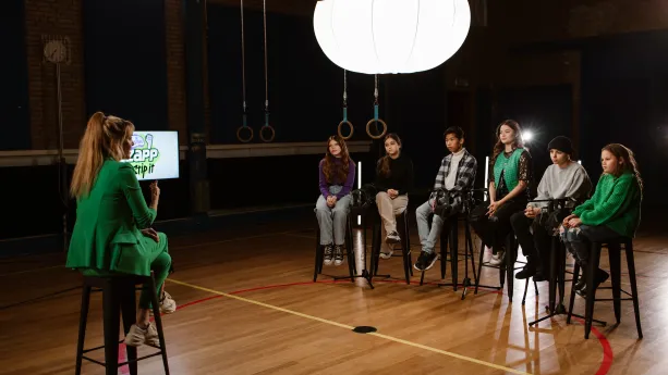 Een foto van zangeres, actrice en presentatrice Stefania Liberakakis die tussen vijf kinderen inzit in het midden van een gymzaal. De kinderen en Stefania zitten op een hoge kruk rechts en links in beeld zit presentator Anne-Mar ook op een hoge kruk.
