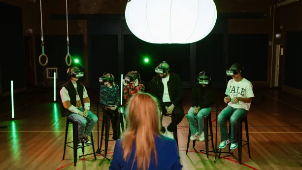 Een foto van rapper en YouTuber Qucee die tussen vijf kinderen inzit in een zaal. De kinderen en Qucee zitten op een hoge kruk en hebben een VR, Virtual Reality, bril op met de tekst "De Pestbril" erop geschreven. De rug van Anne-Mar is vooraan te zien.