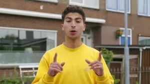 Een foto van Nizar die in een straat staat met huizen op de achtergrond. Hij heeft een gele trui aan en wijst met zijn twee wijsvingers omhoog. Voor het huis direct achter Nizar staat een wit bankje en daarvoor een groene struik.