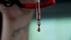 Een foto van de onderkant van een glazen pipet met dikke limonade erin. De pipet zit in een stalen klem. Uit de pipet komt een druppel dikke limonade naar beneden. Dit komt doordat de presentator in het niet-zichtbare deel van de pipet heeft gedrukt.