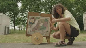 Een foto van presentator Serah die buiten op haar hurken zit voor een houten bord met houten wielen eronder. Op het bord zit een oude landkaart van Nederland geplakt. Serah heeft een t-shirt aan met korte broek en ze draagt sandalen. 
