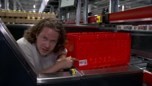 Een foto in een distributiecentrum van een supermarkt voor bestellingen die online geplaatst zijn. Pascal staat achter een rode plastic krat met streepjescode, die voor hem staat op rollende buizen. Een streepjescodescanner ligt ervoor op een stalen rand.