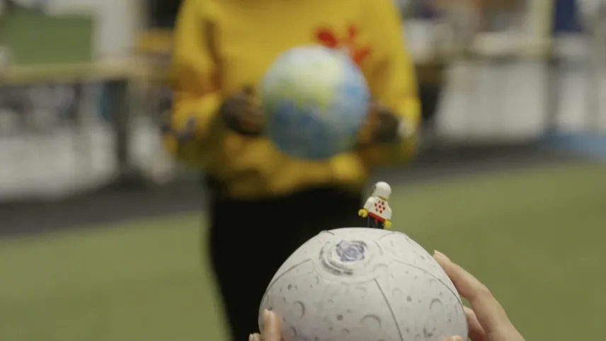 Een foto van twee handen die een opblaasversie van de maan vasthouden. Op de maan staat een LEGO poppetje met een astronautenpak aan. Op de onscherpe achtergrond staat iemand met een opblaasversie van de planeet aarde in zijn of haar handen.