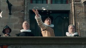 Een foto van een man verkleed als Maurits, de zoon van Willem van Oranje, die muntgeld naar beneden gooit. Hij staat met vier mannen op de verhoogde stoep voor een oud gebouw. Hij heeft een witte kraag om, een lichtbruin vest aan en een zwarte hoed op. 