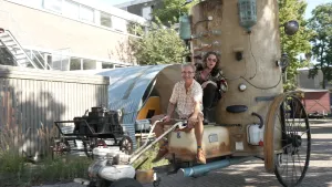 Een foto van Pascal met kunstenaar Olaf Mooij op een auto die Olaf gemaakt heeft en "De Koets" noemt. De auto heeft een bad als zitvlak dat tegen een rechtopstaande hoge, ronde stalen bak, een Silo, aanzit. Olaf houdt het hengsel vast van de motor voorop.