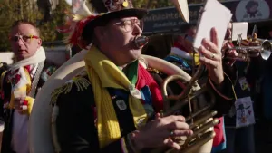 Een foto waarop muzikanten en één carnavalsvierder staan. Groot in beeld is een man in carnavalskleding die het muziekinstrument Tuba bespeeld. Hij kijkt naar bladmuziek in zijn hand tijdens het spelen. De man draagt een hoed met veren en twee sjaals. 