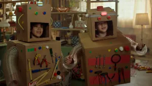 Een foto van Saïd en Anna die verkleed zijn als robot. Ze hebben een kartonnen doos om hun middel waar gereedschap op geplakt zit, een kartonnen doos met een vierkante opening en een lampje erop op hun hoofd en doorzichtige plastic buizen om hun armen.