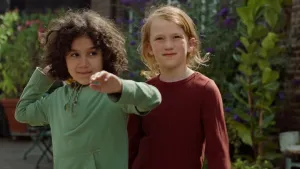 Een foto van Saïd en Anna die buiten staan naast een huis en voor groene struiken en paarse bloemen. Saïd heeft zijn rechterarm naar achteren gebogen en zijn linkerhand naar voren. Hij staat op het punt de boemerang in zijn rechterhand te gaan gooien.