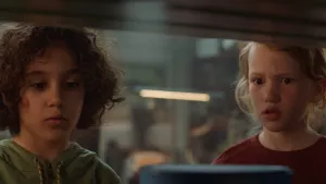 Een foto van Saïd en Anna die door een glazen ruit van een kast kijken.In de kast is nog net onscherp de ronde bovenkant van een blauw verfblik te zien. Saïd en Anna willen de muur van hun hut gaan verven. 