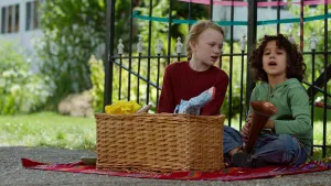 Een foto van Saïd en Anna die op een rood picknickkleed zitten voor een zwart hek. Het kleed ligt op een grindpad. Voor hen op het kleed staat een rieten mand met spullen erin die ingepakt zijn. Saïd heeft een muziekinstrument vast met een hoorn.