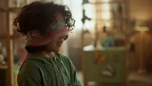 Een foto van Saïd die geblinddoekt is met een sjaal van meerdere kleuren. Saïd heeft een groene trui met capuchon aan. Op de achtergrond is onscherp een groen meubel met een gele en een witte vlinder erop te zien.