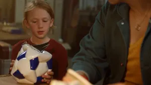 Een foto van Anna die kijkt naar de lekke wit en blauw gekleurde voetbal die ze vastheeft en op een tafel heeft gelegd. Daarnaast zit iemand die niet volledig in beeld is aan de tafel. Op de achtergrond is een donkere en onscherpe kamer te zien.