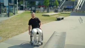 Een foto van Reto, een bewegingswetenschapper, die in een speciale door hem gemaakte rolstoel met voorwielbesturing zit. Reto zit op een plein in Utrecht waar ook twee bankjes staan. Achteraan zijn twee bushokjes, een roltrap en trappen te zien. 