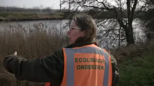 Een foto van een vrouw die een oranje hesje met zilveren strepen aanheeft. Op het hesje staat "Ecologisch onderzoek". De vrouw staat aan een slootkant met riet en een boom ernaast. De vrouw heeft haar handen uit elkaar om de grootte van iets aan te geven.
