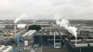 Een foto van een fabriek vanuit de lucht genomen. Op en naast de fabriek staan vier schoorstenen waar witte rookwolken uitkomen. Ook zijn er vier geparkeerde vrachtauto's te zien. Achter de fabriek staan er meer gebouwen op een industrieterrein.
