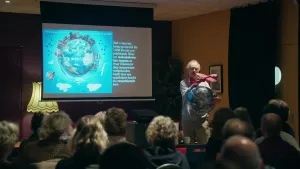 Een foto van een man die een presentatie geeft aan een groep mensen in een zaal. Hij heeft een wereldbol tegen zich aan gedrukt met zijn hand en houdt een rood vliegtuig erboven. Op een scherm  staat "Welcome to the impossible" op een getekende wereldbol.
