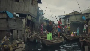 Een foto van een waterweg tussen huizen in Nigeria. Er drijven allemaal houten roeiboten in het water. In zes van de boten staat een man die een lange roeispaan in het water vastheeft. Links staan schuin naar voren hangende huizen van hout.
