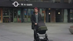 Een foto van een man die buiten loopt met een zwart met groene kinderwagen op een plein in Ijsland. Op de achtergrond is een café zichtbaar, "te & kaffi", wat thee en koffie betekend in het Ijslands. De man draagt een zwarte jas en een rode muts.