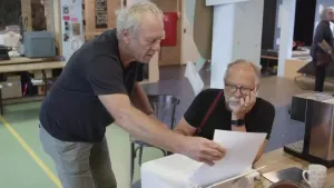 Een foto van twee mannen aan een tafel met een witte printer erop. Eén man zit op een stoel naar de printer te kijken. De andere man staat bij de printer en steekt een wit vel in de staande lade van de printer om daarna te laten zien wat er stuk is.