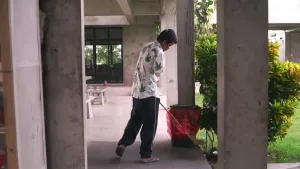 Een foto van een man met een bezem in zijn handen. De man staat op een terras van een huis of gebouw bij een plastic prullenbak met rode zak erin en een groene struik. Hij draagt een trainingsbroek, slippers en een witte blouse met groene bladeren.