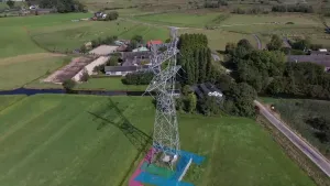 Een foto van een elektriciteitsmast in een groen grasveld. De foto is vanuit de lucht genomen. Van de mast lopen kabels naar andere masten die niet te zien zijn. Er staan bomen en vrijstaande huizen en gebouwen op de foto langs een smalle weg.