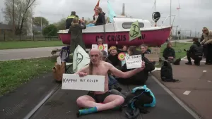 Een foto van een groep mensen op een trambaan die protesteren tegen klimaatverandering. Achteraan staat een rood-witte boot met de tekst "De crisis is nu" erop. Een man zit in zijn onderbroek met twee borden: "de naakte waarheid" en "kappen met kolen".