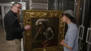 Een foto van een presentator in het depot van het Centraal Museum met medewerker Bart. Ze staan bij het schilderij "Slapende Mars". Het schilderij heeft een opvallende, goudkleurige lijst met wapens, schilden en helmen erop, die aan een ketting zitten.