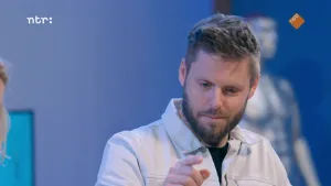 Een foto van prof die met zijn rechterwijsvinger naar voren wijst en naar beneden kijkt. Prof draagt een witte blouse. Links is nog net een randje van het hoofd van Janouk te zien. Rechtsachter prof is een onscherp zilveren beeld van een mens te zien.
