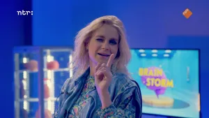 Een foto van Janouk die haar wijsvinger omhoog houdt. Ze heeft haar rechteroog dichtgeknepen, zodat ze een knipoog geeft naar de camera toe. Op de achtergrond staat onscherp een glazen vitrinekast met lichtjes, de voorwerpen erin zijn onherkenbaar. 