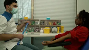 Een foto van een presentator op een stoel. De presentator heeft een arm uitgestrekt en de mouw van de arm opgestroopt.  Links zit een dokter met een band van stof in zijn handen. Op een tafel staan kleine ladenkastjes met onderdelen om te kunnen prikken.