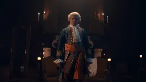Een foto van een man verkleed als stadhouder Willem IV in een donkerer kamer verlicht door kaarsen. Hij draagt een witte pruik, blauw kostuum met goudkleurige knopen en versieringen, een witte sjaal om zijn nek en een rode strik om zijn middel.  