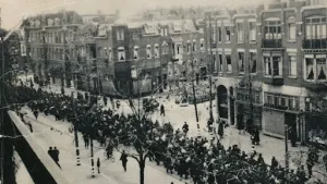 Een zwart-wit foto van de razzia in Rotterdam tijdens de Tweede Wereldoorlog in 1944. Er lopen allemaal mannen dicht op elkaar in het midden van een straat met huizen. Ze hebben van de Duitse bezetter een bevel gehad om zich te melden voor verplicht werk.