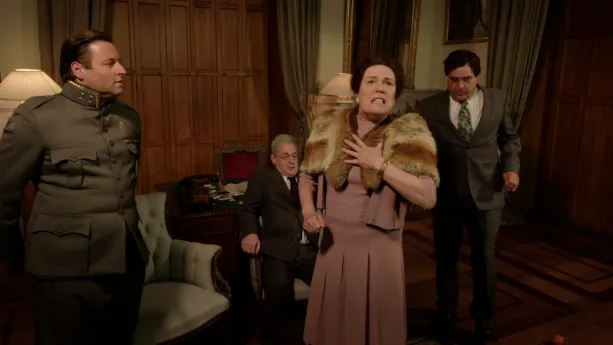 Een foto van koningin Wilhelmina die haar hand op haar borst heeft en omhoog kijkt. De koningin staat in een kamer in paleis Noordeinde. Links naast Wilhelmina staat generaal Winkelman. Achter haar zit een minister en staat een andere minister.