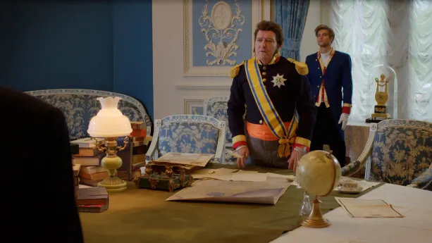 Een foto van koning Willem I achter een grote, ronde tafel. Op de tafel liggen een landkaart en boeken en staan een wereldbol en een kleine lamp. Op de voorgrond is de schouder van een man zichtbaar. Op de achtergrond staat een bediende achter de koning.