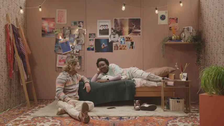 Een foto van Pascal en Tirsa in een kamer. Pascal zit op de grond naast een éénpersoonsbed waar Tirsa op haar zij opligt. Te zien zijn: posters aan de muur van onder andere artiesten, kleding op een houten rek en een wit vloerkleed op een perzisch tapijt.